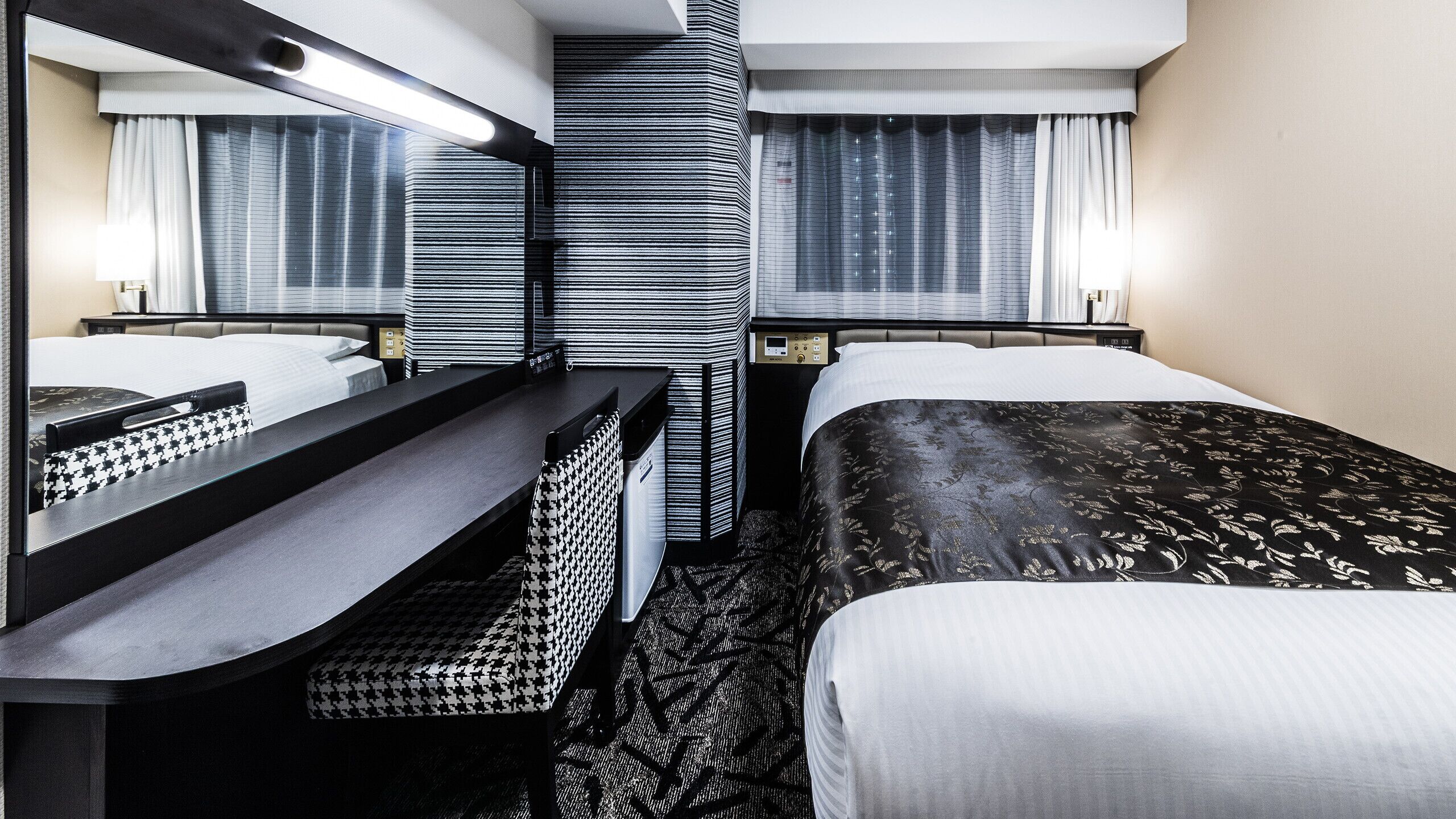 A hotel room with a bed, a desk, a mirror, a control panel, and a window with curtains.