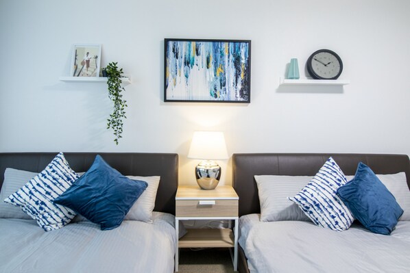 Habitación de hotel con dos camas, una mesita de noche con lámpara, un reloj y un cuadro abstracto enmarcado en la pared.