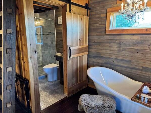 A bathroom with a wooden door, a white bathtub, a fluffy rug, and a wooden shelf with toiletries. - The Seabiscuit at Jayell Ranch (Sevierville)