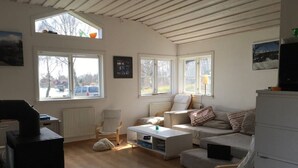 A cozy living room with a sofa, armchairs, a coffee table, and a TV. There are large windows providing natural light and views of the outside. - Cozy and modern cottage by the beach in the south of Sweden (Djursvik)