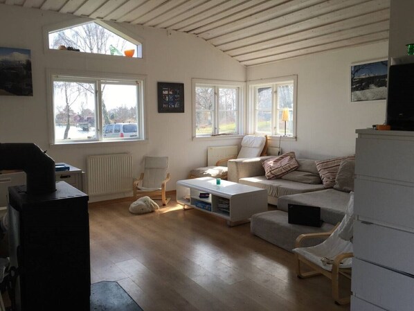 A cozy living room with a sofa, armchairs, a coffee table, and a TV. There are large windows providing natural light and views of the outside.