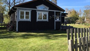 A small, dark wooden building with white trim and two windows, surrounded by a grassy yard and trees. - Cozy and modern cottage by the beach in the south of Sweden (Djursvik)