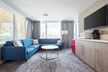 A modern hotel room with a blue sectional sofa, a round coffee table, a flat-screen TV, and a kitchen area with a red refrigerator.. The Cole