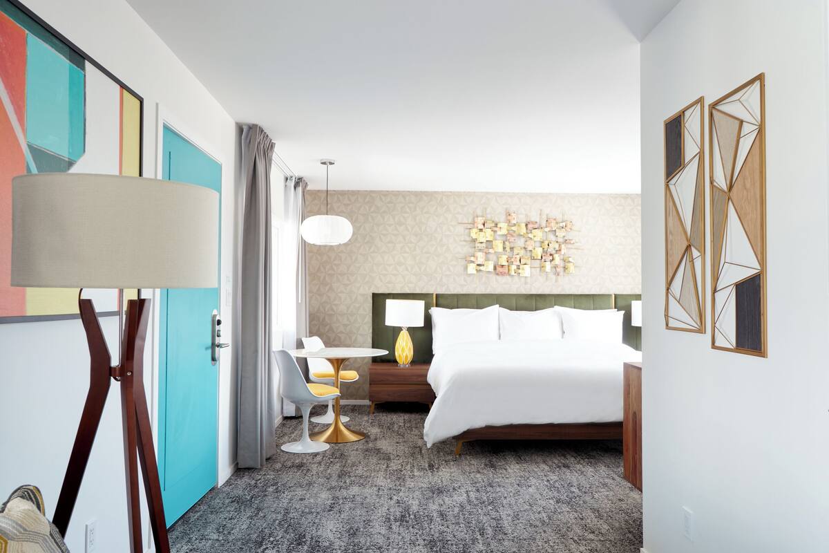 A hotel room with a bed, a desk with a lamp, a chair, and a wall with geometric decor.