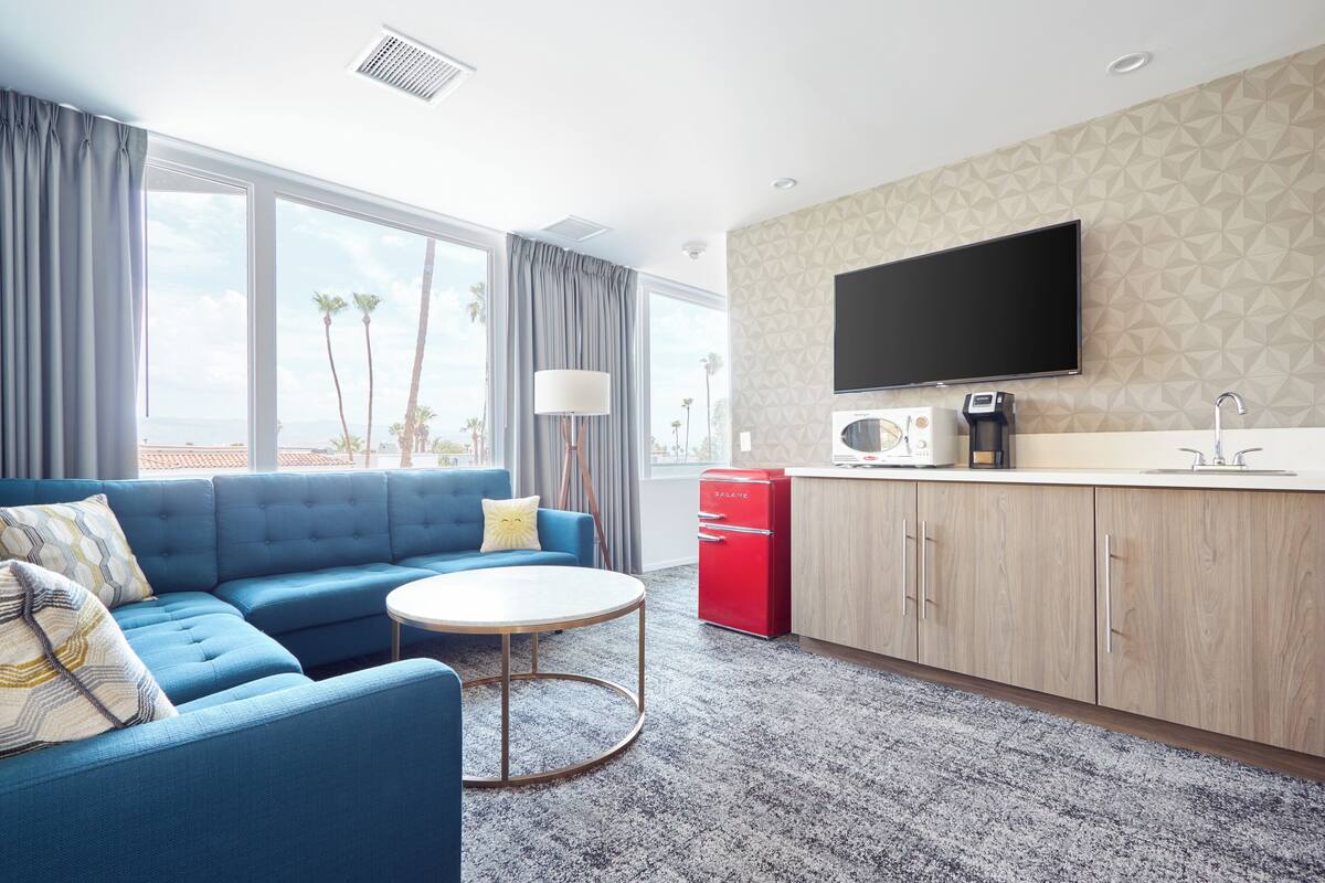 A modern hotel room with a blue sofa, a round coffee table, a flat-screen TV, a kitchenette, and large windows with a view of palm trees.