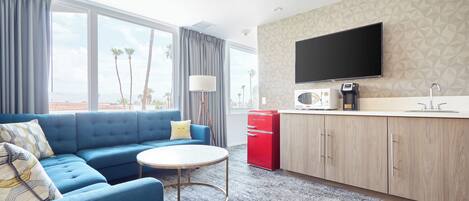 A modern hotel room with a blue sofa, a round coffee table, a flat-screen TV, a kitchenette, and large windows with a view of palm trees.