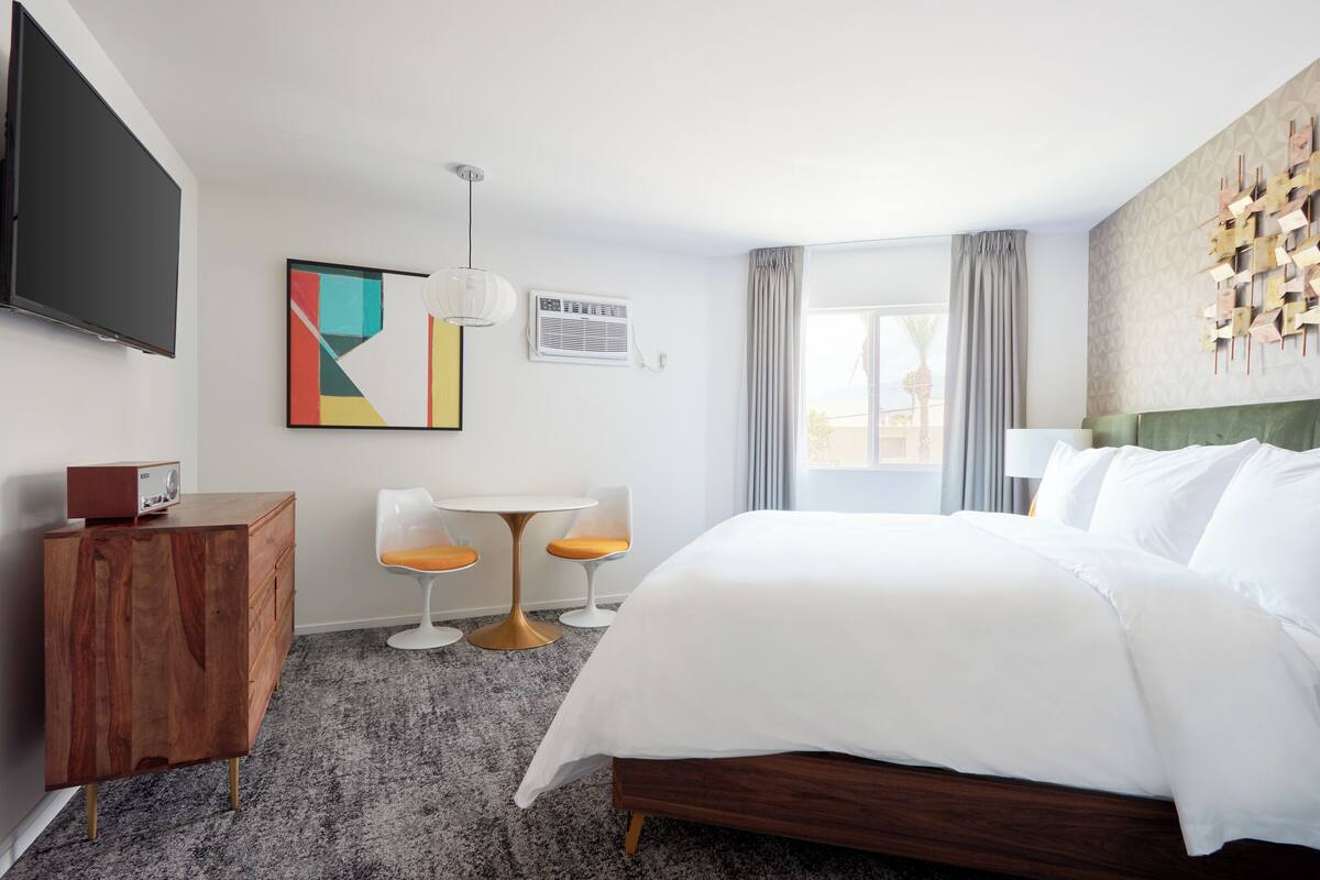 A hotel room with a large bed, a wooden dresser, a television, a small table with two chairs, and a window with curtains.