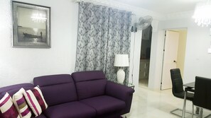 A living room with a purple sofa, a framed picture on the wall, a patterned curtain, a standing lamp, and a dining area with a chair.