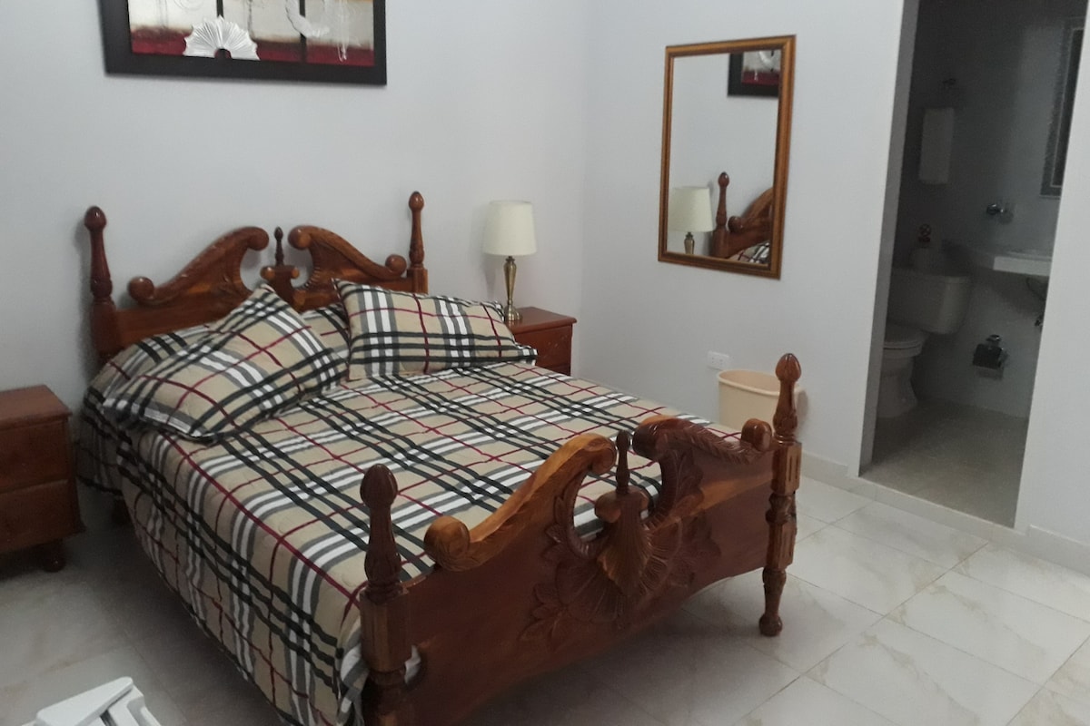 A bedroom with a wooden bed, a nightstand, a mirror, and a bathroom visible through an open door.