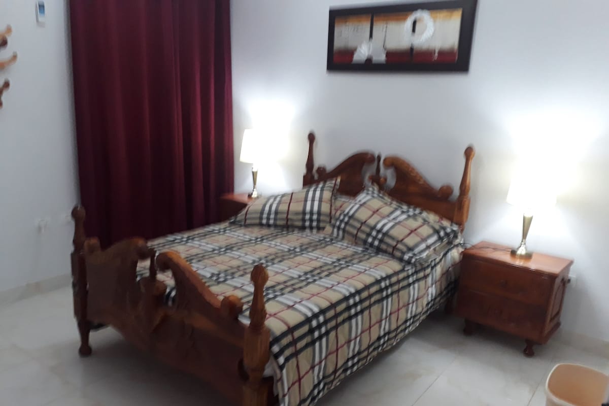 A bed with a plaid bedspread, wooden headboard, two bedside tables with lamps, a red curtain, and a framed picture on the wall.