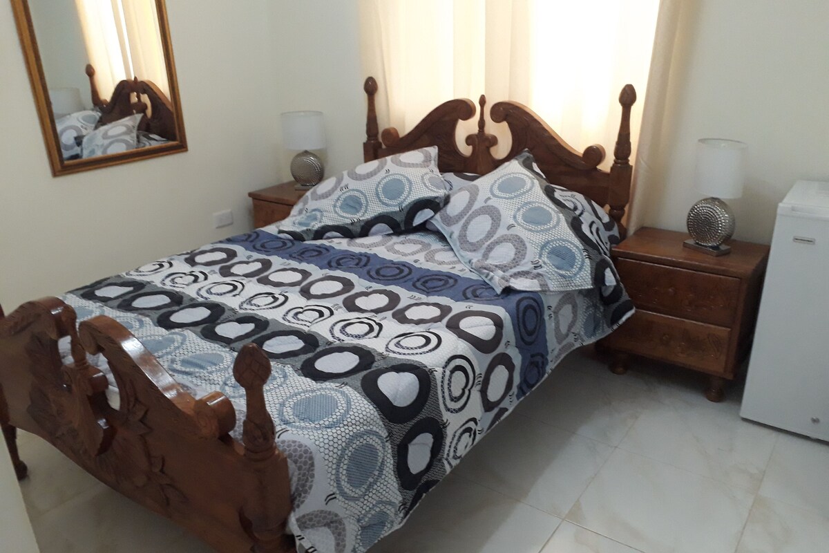 A bed with a patterned bedspread, wooden headboard, two bedside tables with lamps, a mirror, and a small white appliance on the right.