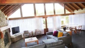 Un salón espacioso con chimenea de piedra, asientos cómodos, un televisor de pantalla plana y ventanas amplias con cortinas traslúcidas.