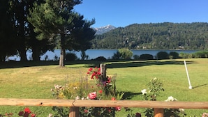Un área con césped, una valla de madera, un árbol grande y una vista al lago y las montañas a lo lejos.