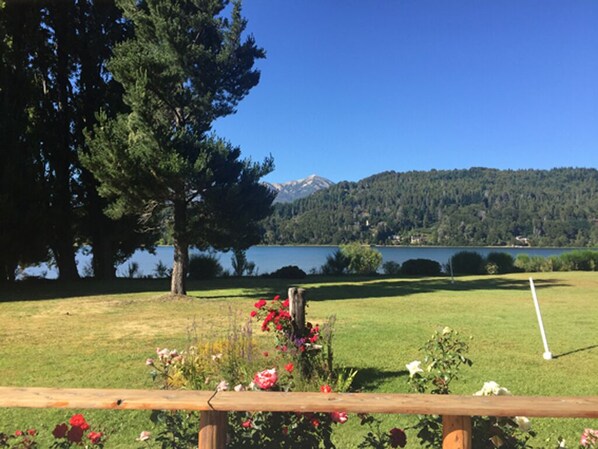 A grassy area with a wooden fence, a large tree, and a view of a lake and mountains in the distance.
