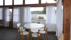 Un espacio comedor al aire libre cubierto con una mesa redonda y seis sillas, ventanas amplias con cortinas blancas y vistas a un paisaje natural.