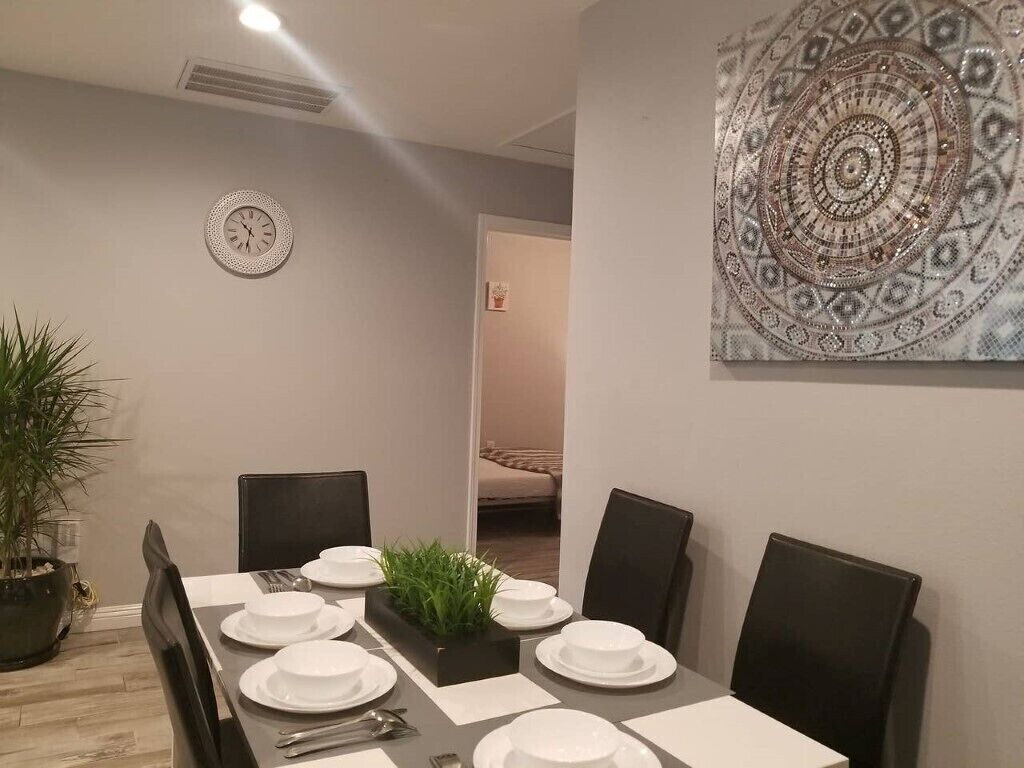 A dining area with a long table set for four, a clock on the wall, and a decorative tapestry.