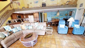 Uma sala de estar ampla com um sofá seccional bege grande, uma mesa de centro redonda e uma poltrona azul. Uma cozinha é visível ao fundo, com armários de madeira e uma área de jantar com mesa e cadeiras.