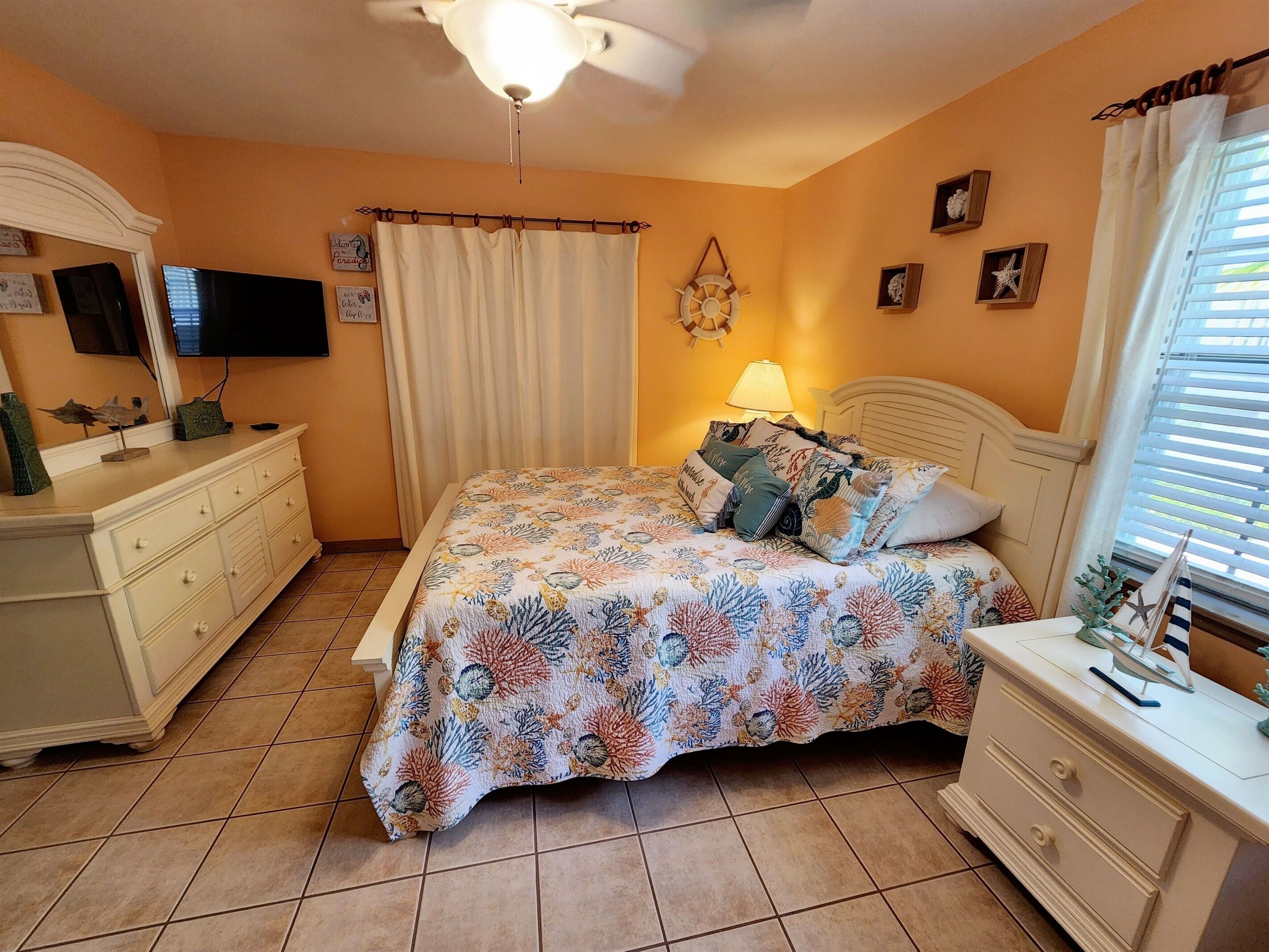 A bedroom with a bed, a nightstand, a dresser, a ceiling fan, a TV, and a window with curtains.