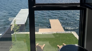 A room with a large window overlooking a lake, a wooden dock, and a grassy area with outdoor furniture.