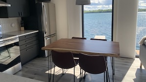 A modern kitchen with dark cabinets, a stainless steel refrigerator, and a wooden dining table with four chairs. A large window offers a view of a lake and distant trees.
