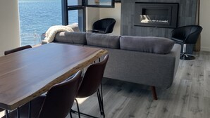 A modern living room with a wooden dining table, grey sofa, and black chairs. A large window offers a view of the water.