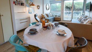 Un espace repas moderne avec une table ronde pour quatre personnes, un canapé avec des coussins et une grande fenêtre donnant sur un paysage verdoyant.