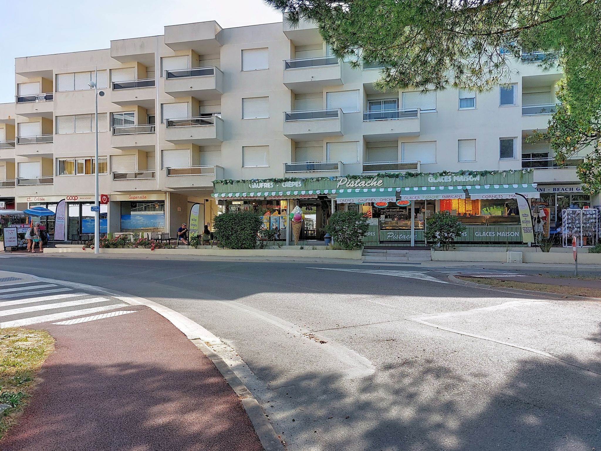 Ein mehrgeschossiges Gebäude mit Balkonen, eine Straße davor und ein kleiner Laden mit einer grünen Markise.