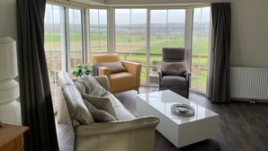 A modern living room with a sofa, armchairs, a coffee table, and large windows offering a view of the countryside.