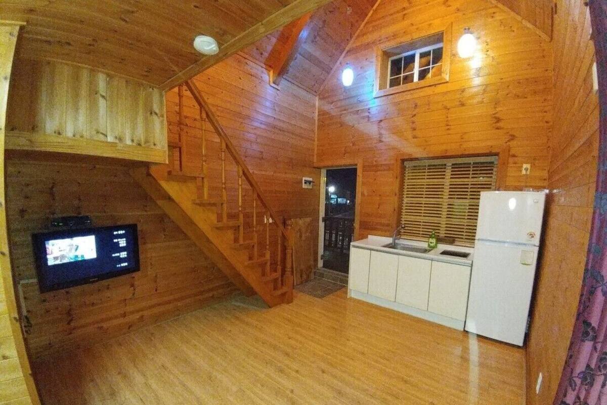 Interior de una cabaña de madera con una zona de cocina, una escalera y un televisor montado en la pared.