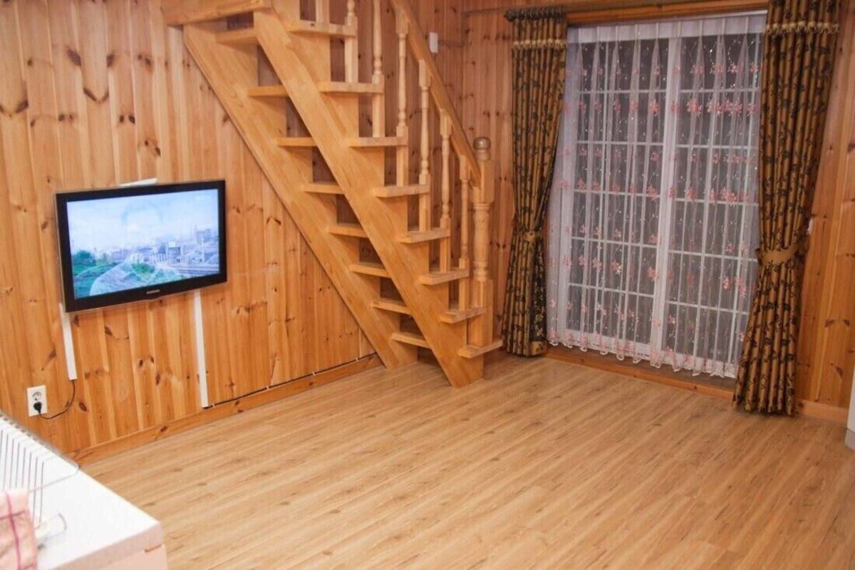 Una habitación con paredes y suelos de madera, una escalera de madera, un televisor montado en la pared y una ventana con cortinas estampadas.