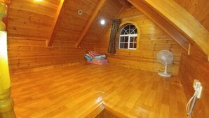 A wooden interior with a sloped ceiling, a window with curtains, and a fan on the floor. - Yangsan Park Pension (Yangsan)