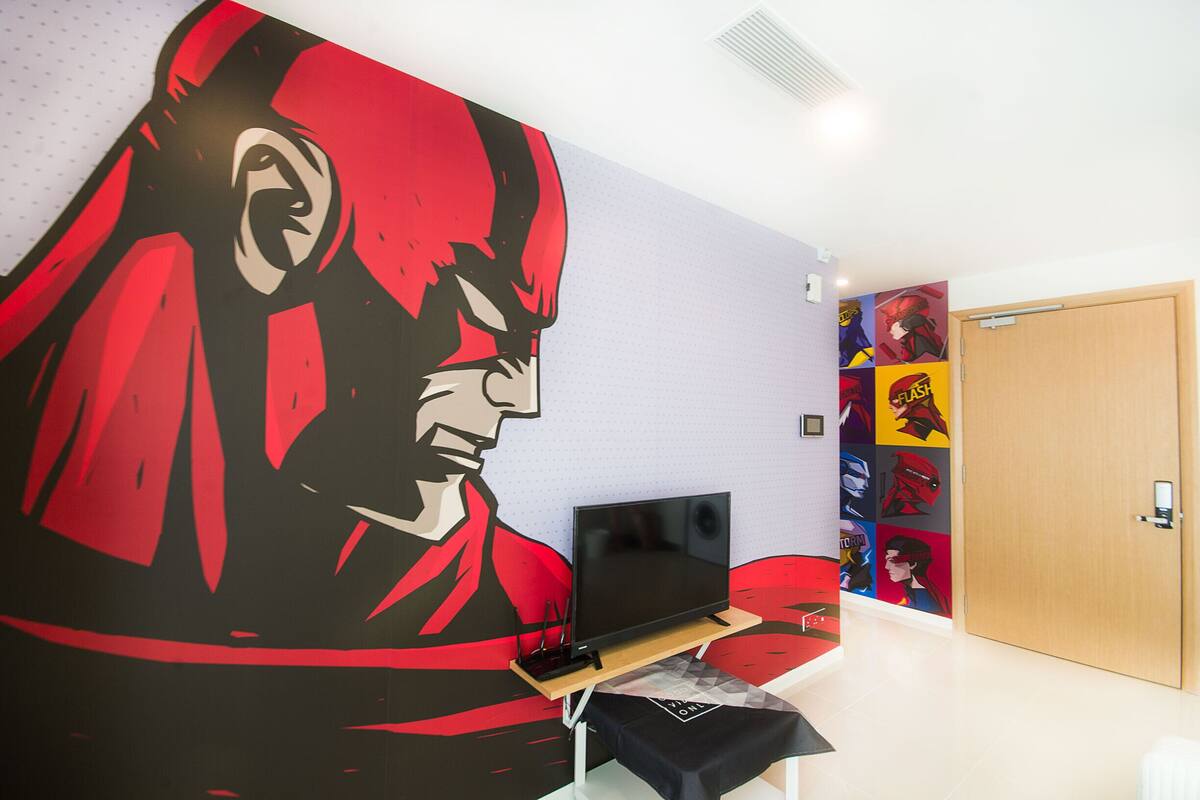 A room with a large wall mural, a flat-screen TV on a stand, and a door with a colorful poster.