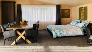 A room with a wooden ceiling, a large bed with patterned bedding, a dining table with chairs, and a window with sheer curtains.