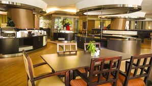 A modern hotel lobby with a central dining area featuring a wooden table and chairs, a buffet counter, and a large decorative plant. - Bela Hotel (Ternate)