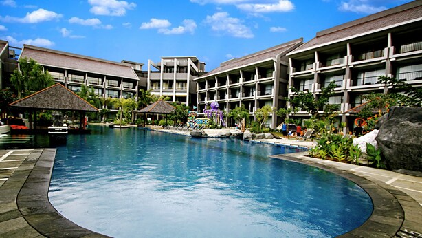 A large swimming pool surrounded by multi-story buildings with balconies, a small waterfall feature, and tropical landscaping. - Bela Hotel (Ternate)