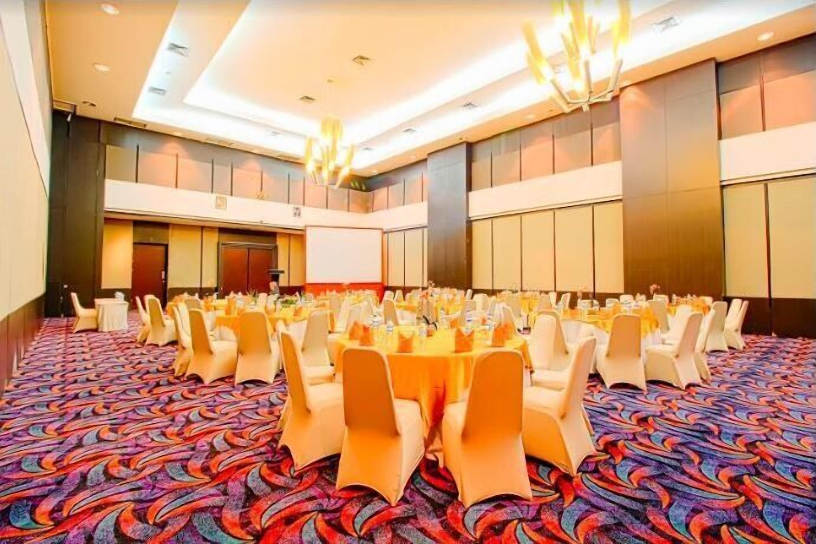 Un salón de banquetes con mesas redondas, sillas blancas y una alfombra colorida.