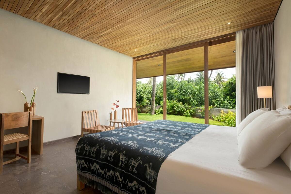 A bedroom with a bed, a desk, chairs, a TV, and a large window with a view of greenery.