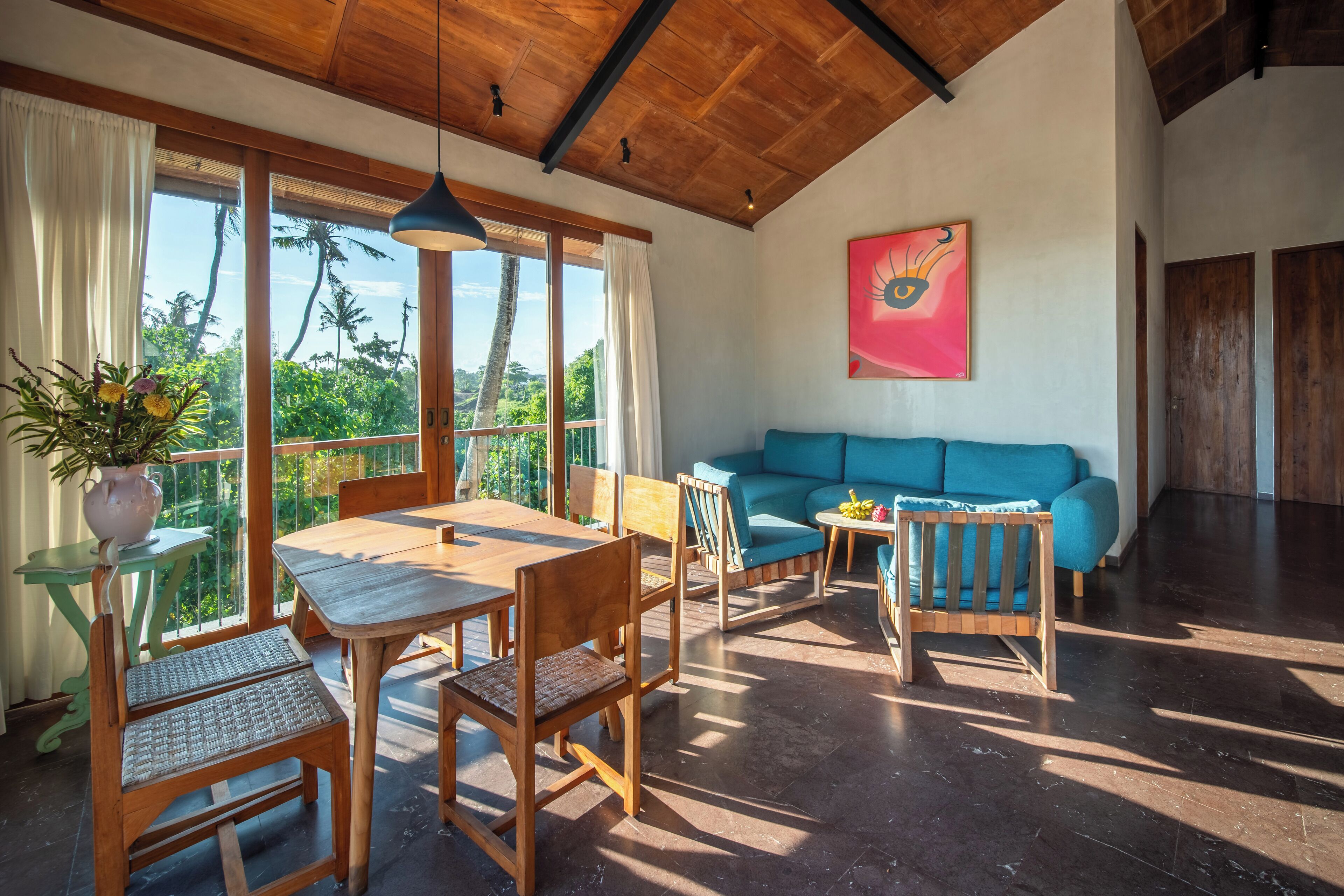 Un'ampia zona giorno con soffitto in legno, un divano blu, un tavolo da pranzo con sedie e ampie finestre che offrono vista su palme.