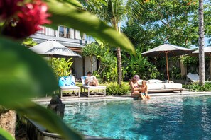 A pool area with lounge chairs and umbrellas, surrounded by tropical plants and a building in the background. - MASMARA Resort Canggu (Pererenan)