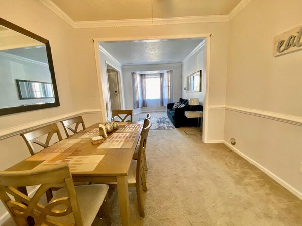 A dining area with a long table and chairs, a mirror on the wall, and a hallway leading to a living room with a couch and a TV.