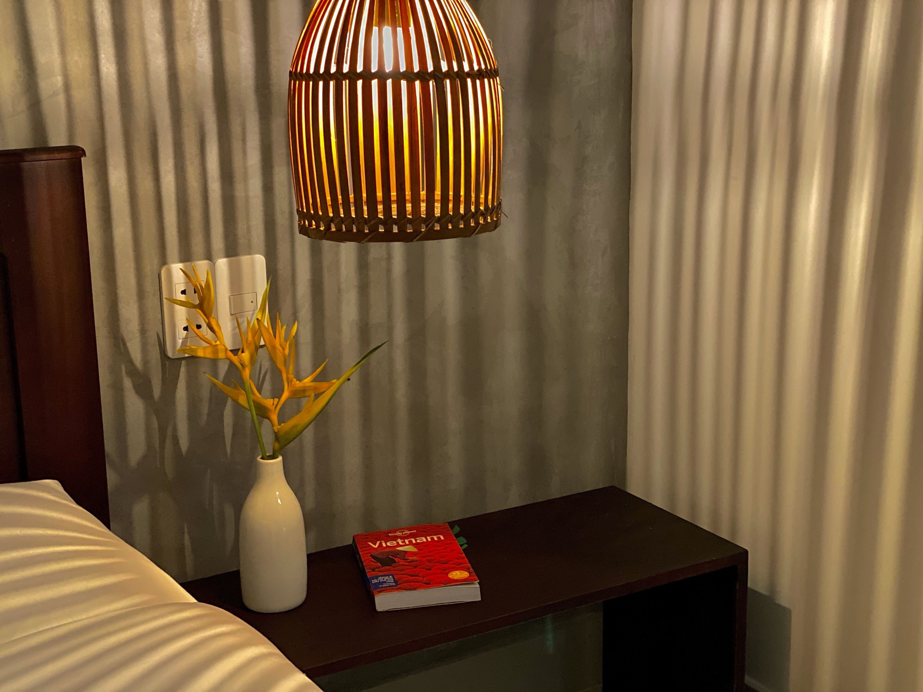 A hotel room with a bedside table, a white vase with a plant, a book titled 'Vietnam', and a hanging lamp.