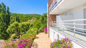 Un bâtiment moderne à la façade en pierre, doté d’un balcon avec une rambarde en verre, et d’un chemin bordé de fleurs roses qui mène à une colline verdoyante.