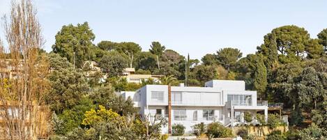A modern two-story house with a flat roof, surrounded by dense greenery and trees.