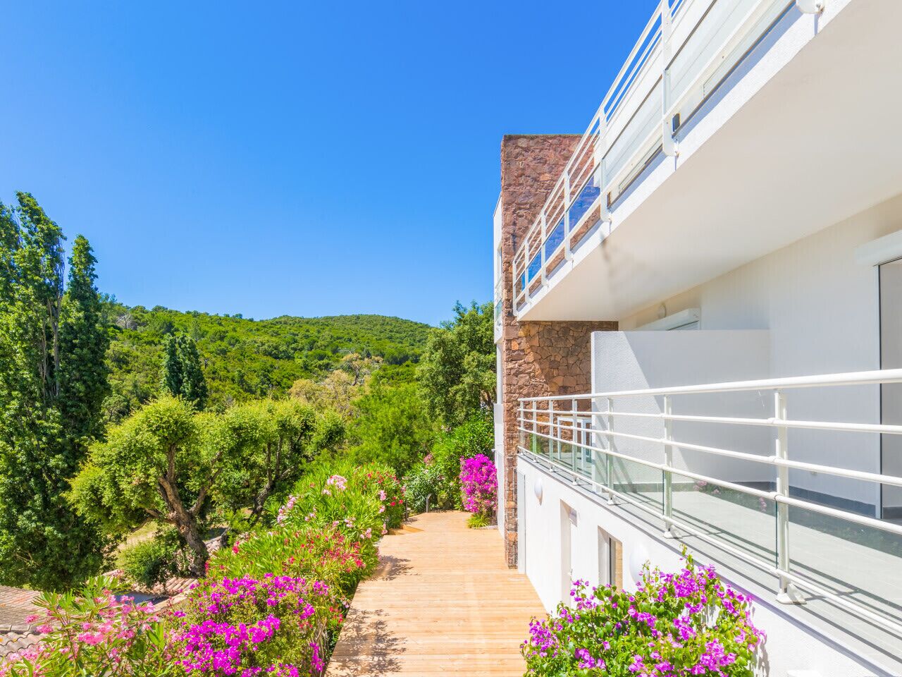 Een modern gebouw met een stenen gevel, een balkon met glazen hekwerk en een pad bezaaid met roze bloemen dat naar een weelderige groene heuvel leidt.