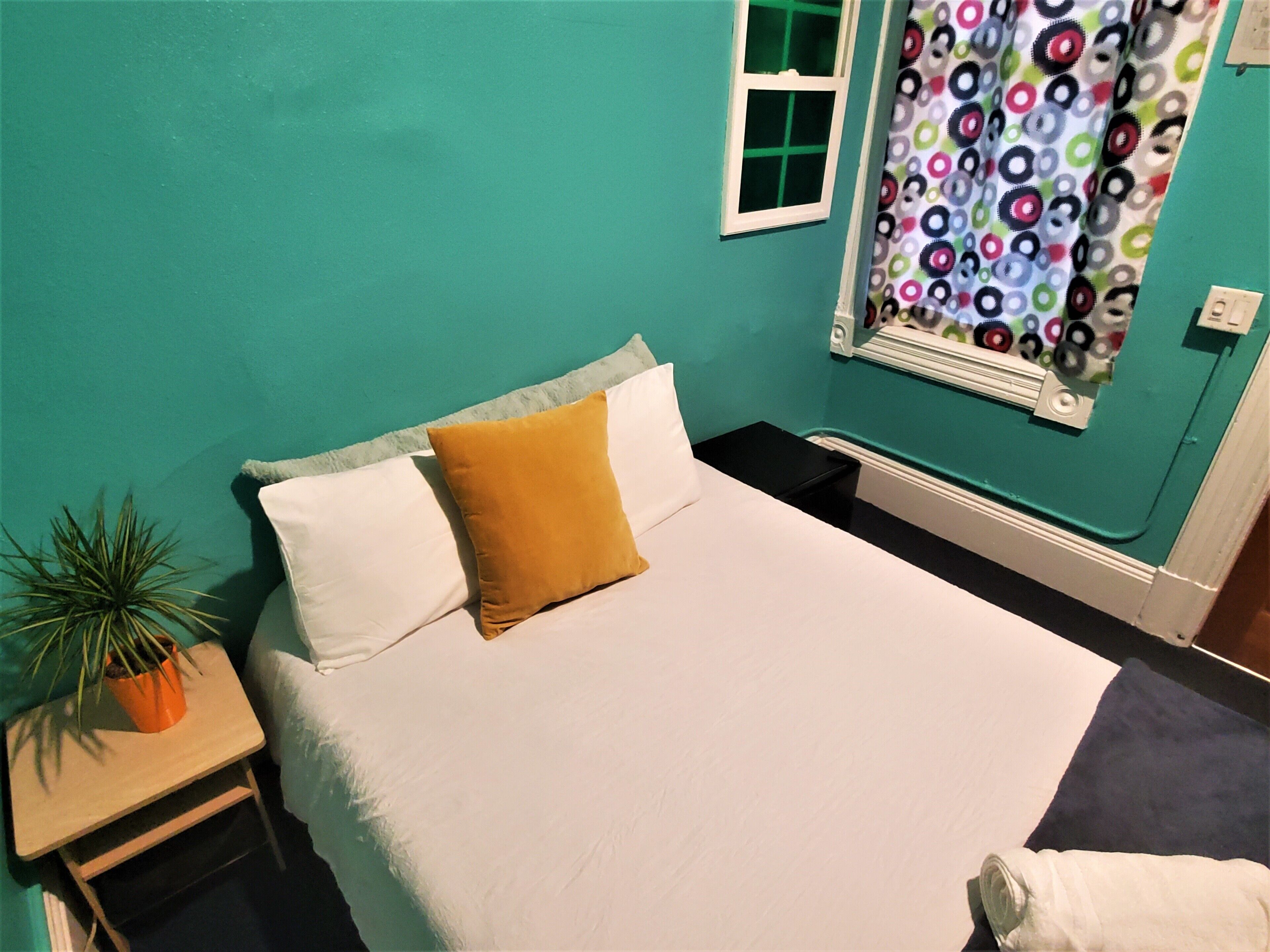 A room with teal walls, a bed with white bedding and a mustard-colored pillow, a small wooden nightstand with a potted plant, a window with a colorful curtain, and a white radiator.