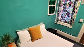A room with teal walls, a bed with white bedding and a mustard-colored pillow, a small wooden nightstand with a potted plant, a window with a colorful curtain, and a white radiator.