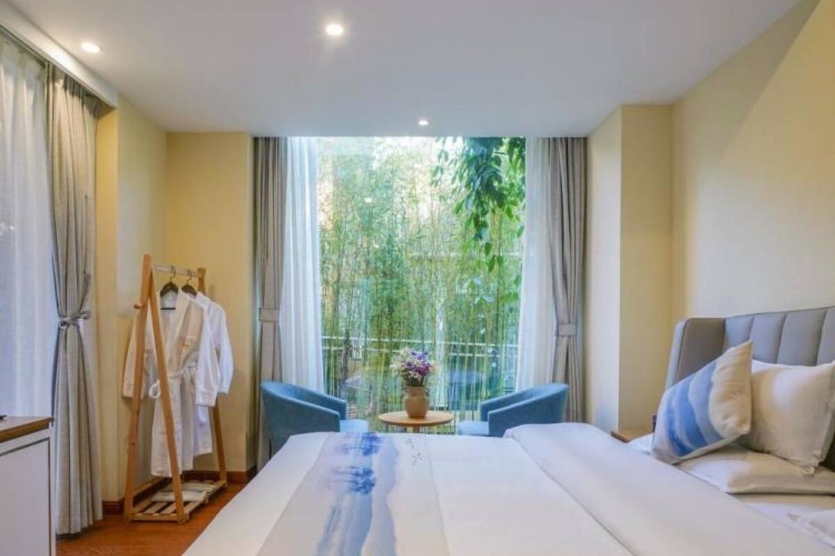 A hotel room with a large bed, a desk, and a chair. There is a window with a view of greenery outside.