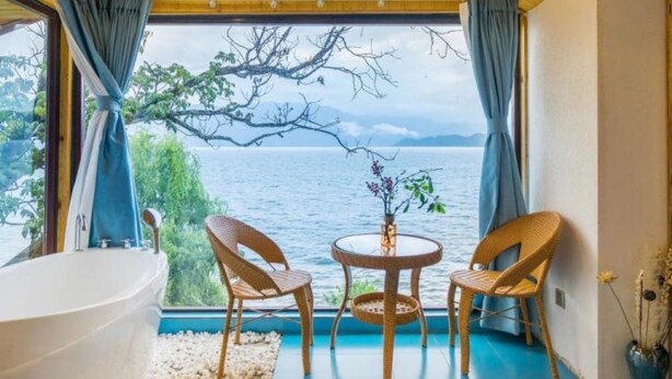 Una habitación con techo de madera, una bañera grande, dos sillones de mimbre, una pequeña mesa redonda y vistas a un cuerpo de agua.