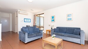 Sala de estar com dois sofás azuis, uma mesa de centro de madeira e uma mesinha com abajur. Há quadros nas paredes e uma porta que dá para outro cômodo.