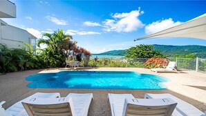 Uma área de piscina com cadeiras de descanso brancas, piscina e vista para o mar e colinas.